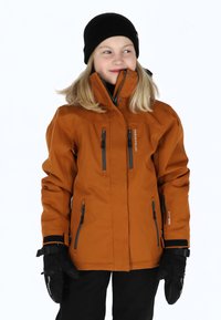 Orange Outdoor-Jacke mit schwarzen Reißverschlüssen und Logo. Verfügt über einen hohen Kragen und mehrere Reißverschlusstaschen. Schwarze Strickmütze und Handschuhe sind getragen.