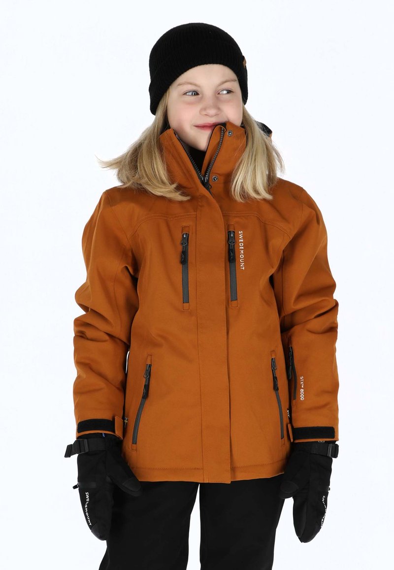 Orange Outdoor-Jacke mit schwarzen Reißverschlüssen und Logo. Verfügt über einen hohen Kragen und mehrere Reißverschlusstaschen. Schwarze Strickmütze und Handschuhe sind getragen.