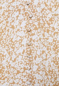 Tissu à motif floral dans des tons beige et blanc avec une fermeture à boutons sur le devant ; texture douce et petits détails de fleurs délicates tout au long.