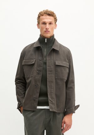 Homme portant une veste boutonnée gris foncé sur un pull à col montant zippé et un pantalon foncé, debout devant un fond blanc uni.