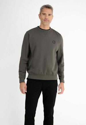 Mand iført en grøn sweatshirt med rund halsudskæring og lille logo på brystet og sorte bukser, stående foran en hvid baggrund.
