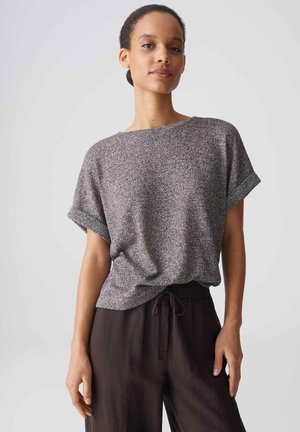 Vrouw met donker haar in een lage knot, een gestructureerde grijze top met korte mouwen en losse zwarte broek, staand voor een effen achtergrond.