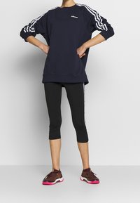 Sudadera navy con diseño de tres franjas blancas en las mangas, combinada con leggins capri negros con franjas laterales; zapatillas deportivas marrones y rosas.