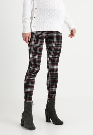 Jersey blanco texturizado con botones laterales, pantalones ajustados de cuadros negros y rojos, y botines de ante negro con tacón en una persona de pie.