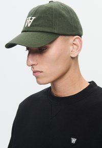 Groene katoenen pet met een geborduurd wit "W"-logo, met een gebogen rand en een verstelbare band aan de achterkant. Draagt met een zwart sweatshirt.