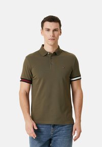 Polo de color verde oliva hecho de algodón, con una abertura de dos botones, mangas cortas con rayas rojas y blancas, y un pequeño detalle de logo.