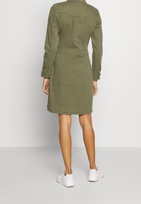 Vestido de algodón verde oliva, a la altura de la rodilla, con cintura ajustada y mangas largas. Cuenta con puños abotonados y bolsillos laterales. Llevado con zapatillas blancas.