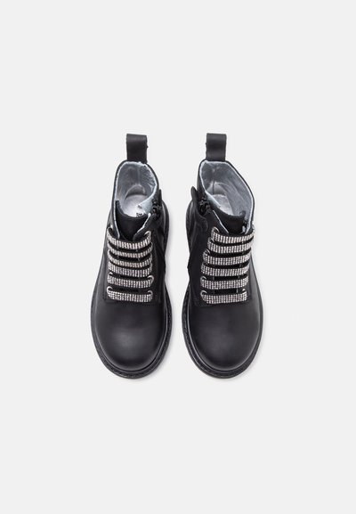 Paire de bottines noires en cuir avec des lacets élastiques ornés de strass et des fermetures éclair latérales, vues de dessus sur un fond clair.