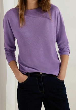 Femme portant un pull violet à manches longues, les mains dans les poches d'un jean bleu foncé, debout devant un fond clair uni.