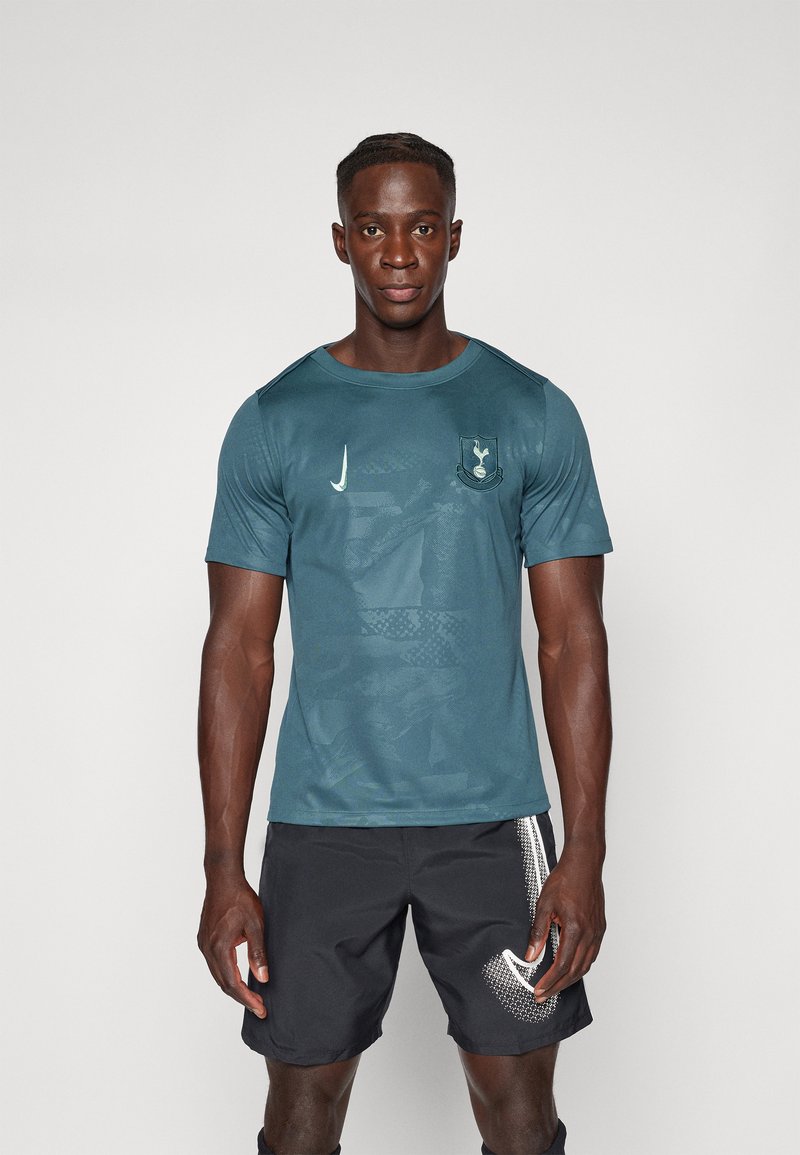Camiseta atlética verde-azul de manga corta, con un logo del equipo y acentos texturizados. Combinada con pantalones cortos negros y un diseño blanco notable.
