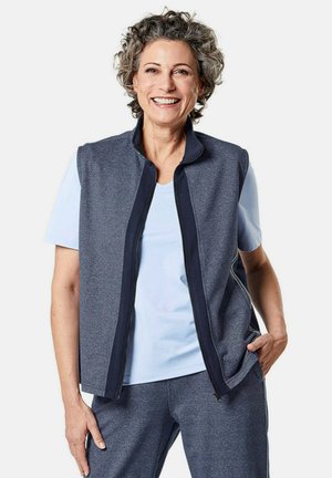 Gilet zippé gris avec bordures bleu marine, porté sur une chemise à manches courtes bleu clair. Tissu doux avec une coupe décontractée. Le modèle se tient avec une main dans la poche.