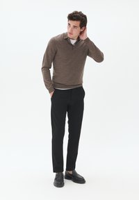 Suéter marrón de punto con cuello y cremallera, combinado con pantalones negros de corte ajustado y mocasines negros, de pie contra un fondo blanco.