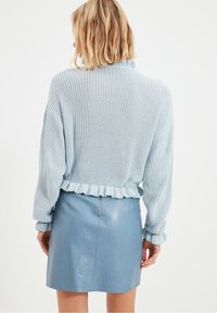 Blondynka odwrócona tyłem, ubrana w jasnoniebieski sweter dziany z falbaniastym dołem i mankietami oraz niebieską skórzaną mini spódniczkę z zamkiem z tyłu.