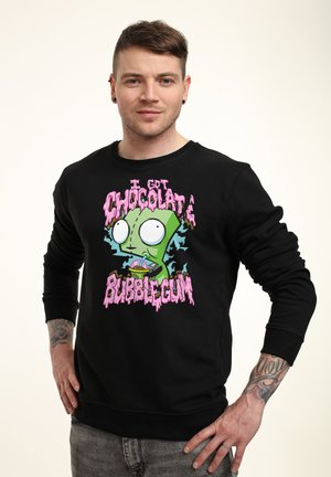 Uomo con felpa nera con personaggio verde da cartone animato e testo rosa "Ho la gomma da masticare al cioccolato", in piedi con le mani sui fianchi.