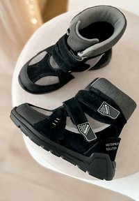 Czarne i szare wodoodporne buty z solidną gumową podeszwą, z rzepami i siateczkowym wykończeniem, z teksturowaną powierzchnią i odblaskowymi akcentami.