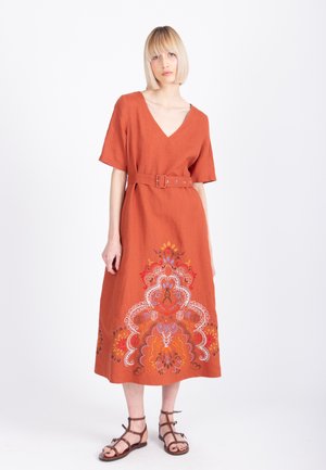 Blondynka stojąca w rdzawo-pomarańczowej sukience z paskiem, ozdobionej wielokolorową kwiatową haftem przy krawędzi i brązowych sandałach na ramiączkach.