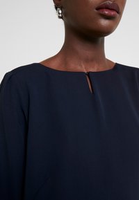 Navyblaues Kleid mit Rundhalsausschnitt und Schlitzdetail, aus glattem Stoff gefertigt, mit genähten Akzenten an den Schultern.