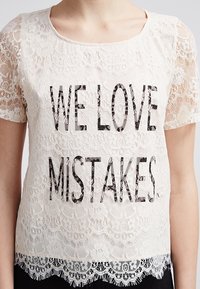 Top de encaje en crema con mangas cortas, que presenta un estampado gráfico negro "NOS ENCANTAN LOS ERRORES". Textura delicada de encaje con dobladillo festoneado.
