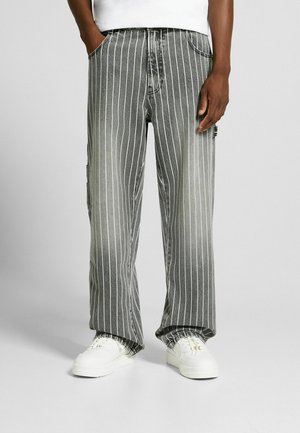 Vaqueros de denim a rayas en negro y gris con un corte holgado, que presentan rayas verticales blancas y bolsillos. Combinados con zapatillas blancas.