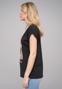 T-shirt en coton noir à manches enroulées, arborant un design graphique coloré sur le devant. Associé à un jean bleu clair, coupe standard.