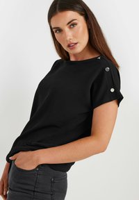 Black short-sleeve top with a round neckline, featuring silver button accents on the shoulders and a textured fabric. Paired with dark gray pants.