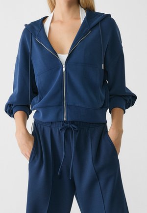 Kobieta ubrana w granatową bluzę z zamkiem na przód oraz pasujące spodnie z sznurkiem, z rękami w kieszeniach i białą bluzką pod spodem.