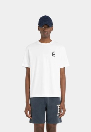 Homme portant un t-shirt blanc avec un logo noir sur la poitrine, un short bleu marine avec un logo blanc, et une casquette de baseball bleu marine, debout face à l'avant.