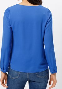 Blaue Langarmbluse aus glattem Stoff, mit Rundhalsausschnitt und geknöpften Bündchen, kombiniert mit eng anliegenden Jeans.