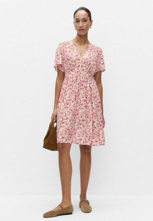 Kobieta w różowo-białej sukience w kwiaty do kolan, trzyma brązową zamszową torebkę, ma na nogach brązowe płaskie buty i złote kolczyki koła.