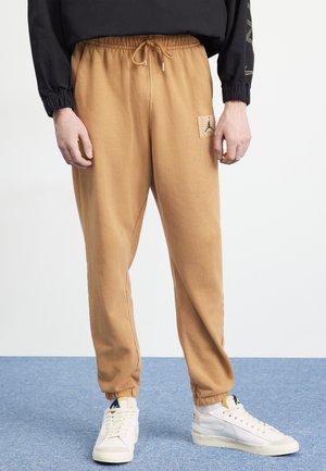Person, die beigefarbene Joggerhosen mit Kordelzug und elastischen Bündchen trägt, mit einem aufgenähten Logo am Oberschenkel, weißen Sneakern und einem schwarzen Langarmshirt.