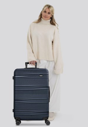 Giovane donna in un maglione chiaro e pantaloni in piedi con una valigia nera texture con manico estensibile e ruote girevoli.