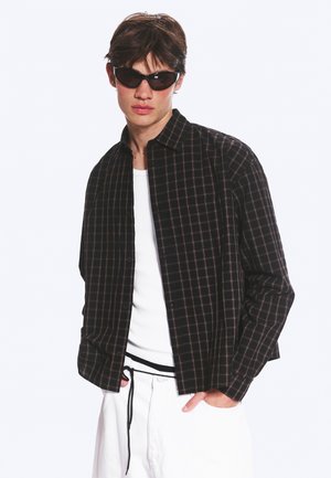 Jeune homme portant une chemise noire à carreaux sur un t-shirt blanc, un pantalon blanc avec cordon et des lunettes de soleil foncées, posant avec une main dans la poche.
