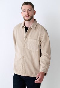 Beige corduroy jacket with a collared neck, buttoned front, and chest pocket. Features vertical ribbed texture and a relaxed fit.