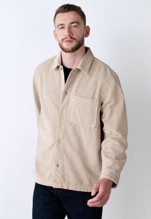 Beige corduroy jacket with a collared neck, buttoned front, and chest pocket. Features vertical ribbed texture and a relaxed fit.