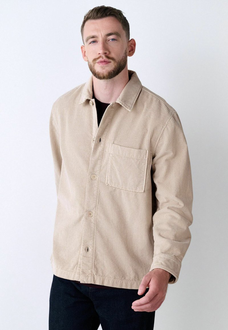 Beige corduroy jacket with a collared neck, buttoned front, and chest pocket. Features vertical ribbed texture and a relaxed fit.