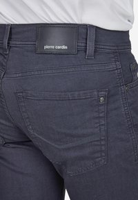 Dunkelblaue Denim-Jeans zeichnen sich durch eine glatte Textur aus, haben ein rechteckiges gesticktes Etikett am Bund mit "Pierre Cardin" und zwei Gesäßtaschen.