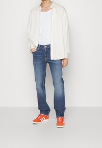 Camisa blanca de botones sobre una camiseta blanca, combinada con unos jeans de mezclilla azules y zapatillas de ante naranjas con rayas blancas.