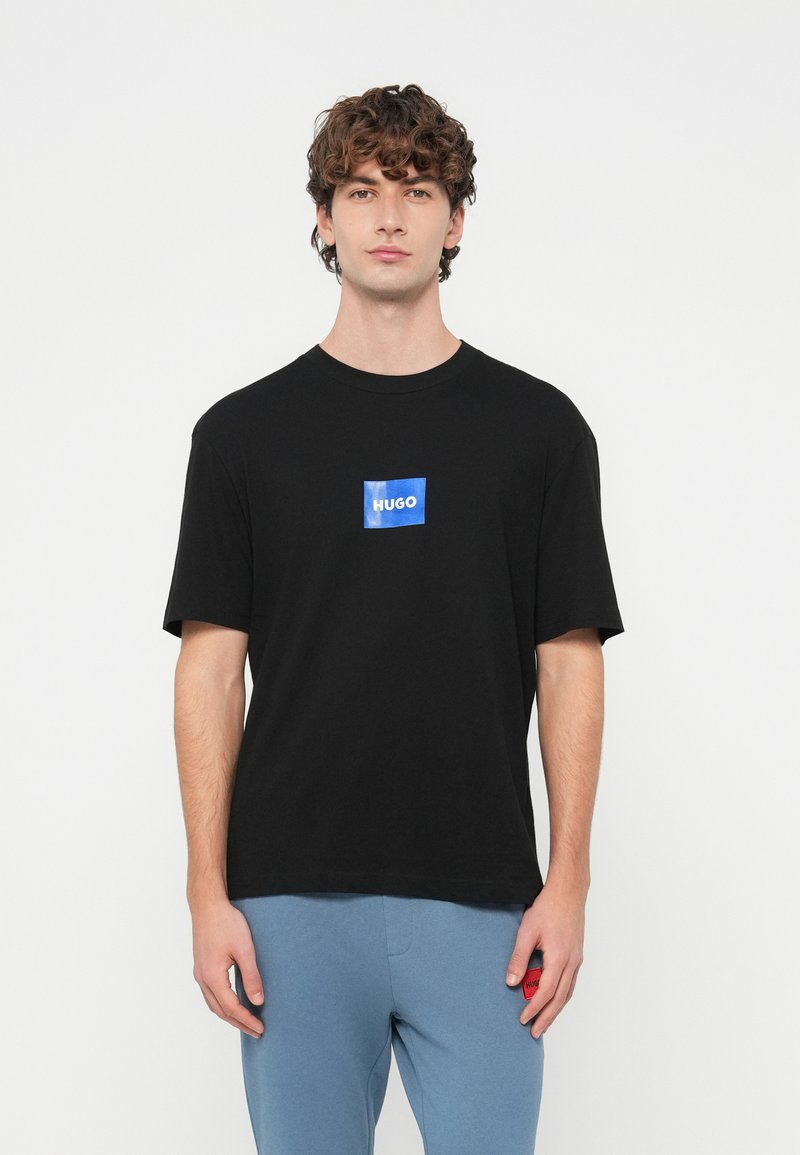 Juoda medvilninė marškinėliai su mėlynu stačiakampiu logotipu, kuriame baltomis raidėmis parašyta „HUGO“, trumpomis rankovėmis ir laisvo silueto.