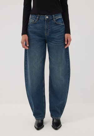 Person trägt weite blaue Jeans, ein schwarzes Langarmoberteil und schwarze spitze Stiefel und steht vor einem schlichten Hintergrund.
