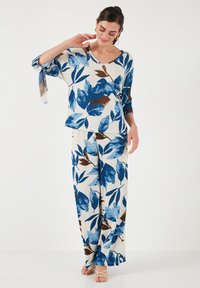 Completo floreale a due pezzi composto da una blusa larga e pantaloni a gamba larga. Predominante bianco con motivi di foglie blu e marroni. Texture morbida.