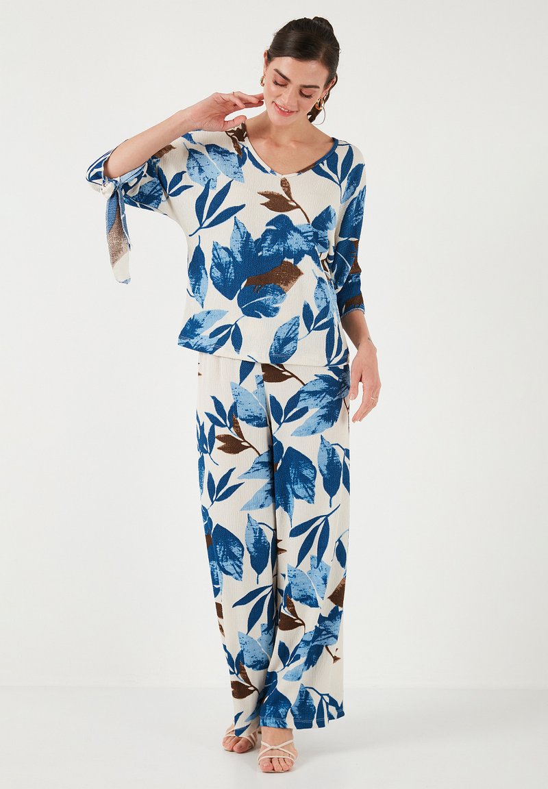 Completo floreale a due pezzi composto da una blusa larga e pantaloni a gamba larga. Predominante bianco con motivi di foglie blu e marroni. Texture morbida.