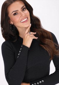 Maglione nero a costine con maniche lunghe, caratterizzato da bottoni decorativi in argento lungo i polsini. I capelli ondulati del modello e gli accessori sono visibili.