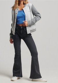 Blouson aviateur en maille gris et gris clair avec poignets rayés, porté sur un débardeur bleu, assorti à un jean évasé foncé et des baskets blanches.