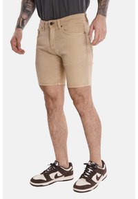 Shorts in denim beige con un design aderente, dotati di tasche frontali, chiusura con bottone e cuciture a vista. Indossati con sneakers marroni e bianche.