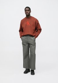 Hnedá pletená mikina s červeným logom, kombinovaná so širokými olivovozelenými nohavicami a čiernymi topánkami na šnurovanie. Hladká textúra, voľný strih.