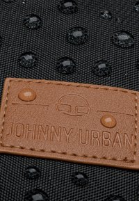 Fekete textúrált anyag, amely emelt kör mintákat és vízcseppeket tartalmaz. Barna bőr címke, amelyre a "JOHNNY URBAN" felirat van gravírozva.