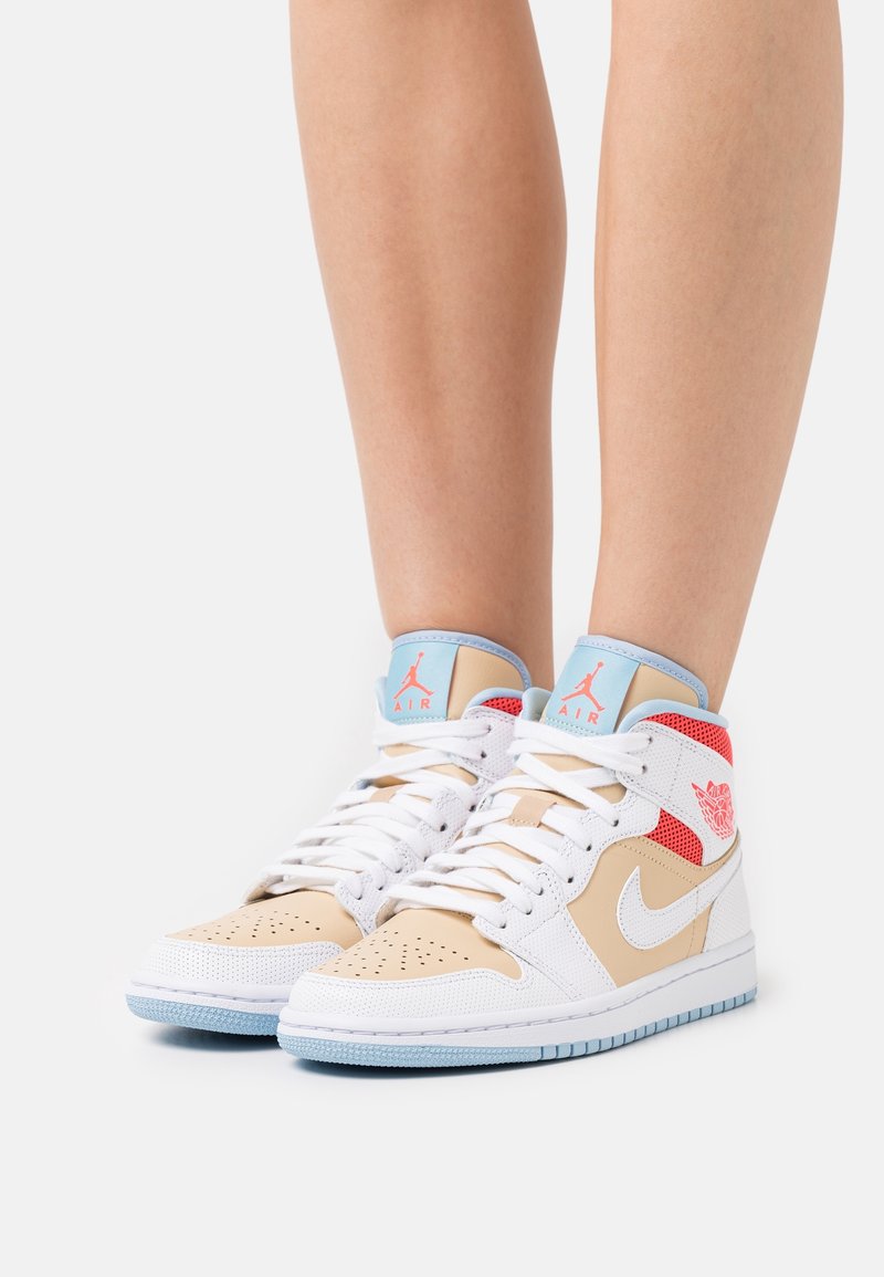 Jambes portant des baskets montantes Nike Air beige, blanches et rouges avec des lacets blancs et des semelles bleues, debout sur un fond clair.