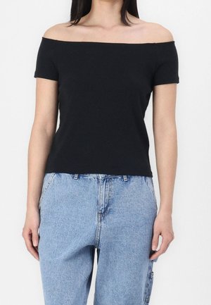 Kobieta ubrana w czarny top z odkrytymi ramionami i krótkimi rękawami oraz jasnoniebieskie spodnie jeansowe z wysokim stanem, stojąca na jednolitym tle.