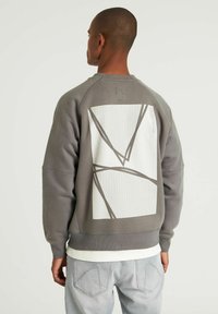 Grå sweatshirts med et hvidt grafik på ryggen, der har geometriske linjer og et tekstureret gittermønster. Ribbet manchetter og kant.