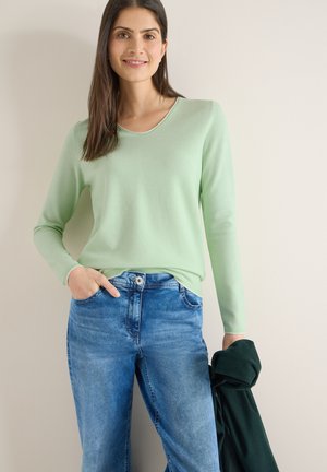 Nainen hymyilee, pukeutuneena vaaleanvihreään pitkähihainen paitaan ja sinisiin farkkuihin, pitäen vasemmassa kädessään tummanvihreää takkia.